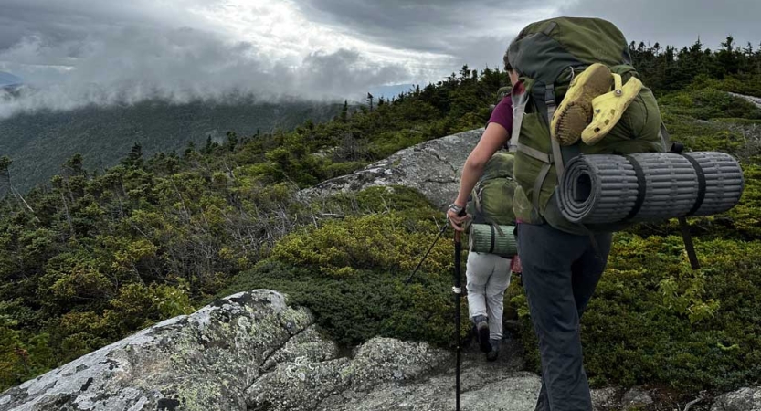 Two students wearing backpacks make their way across a green and rocky landscape. 